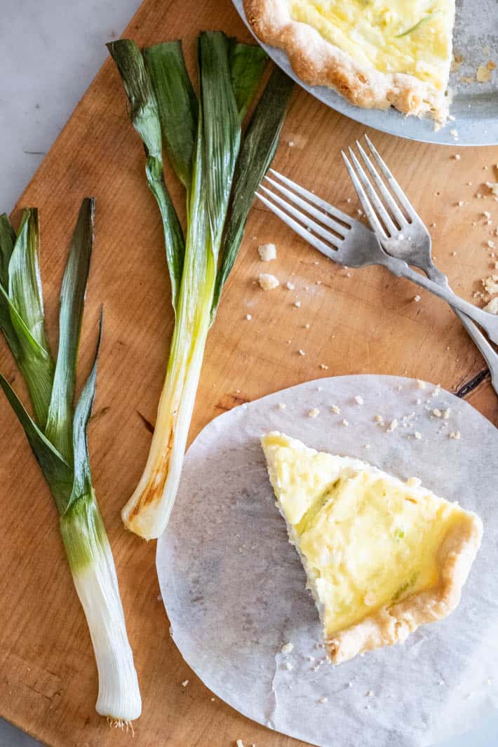 Single slice of leek quiche on parchment paper, with roasted leeks set on a wood serving board. Vintage silver forks crossed over each other above the slice of quiche.