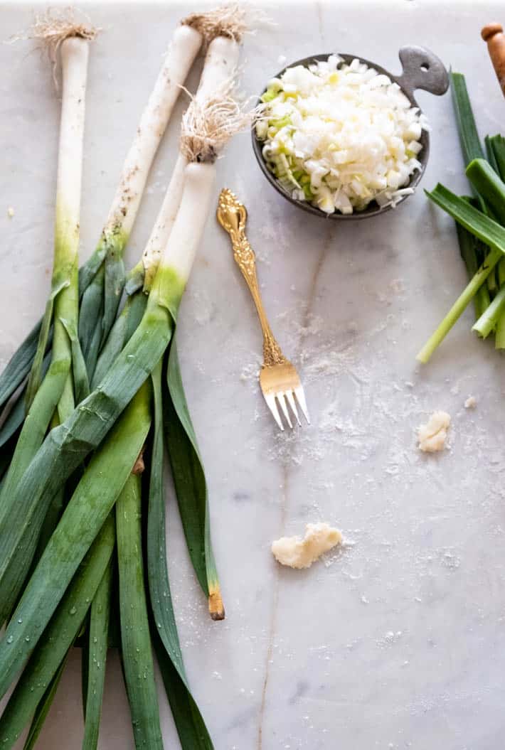 Freshly dug leeks on a marble counter, some prepped and sliced in a measuring cup. Gold fork lays alongside.