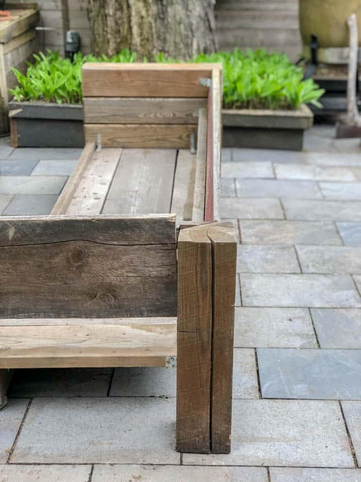 DIY outdoor wooden Restoration Hardware sectional sofa without cushions in front of greenery and large tree on interlocking stone tiles. 