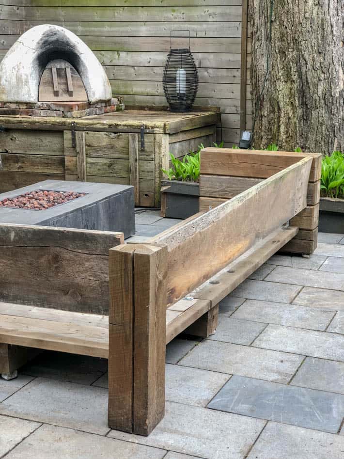 DIY outdoor wooden Restoration Hardware sectional sofa without cushions in front of unlit outdoor fire pit and pizza oven. Outdoor lantern sits beside pizza oven with large tree to the right.