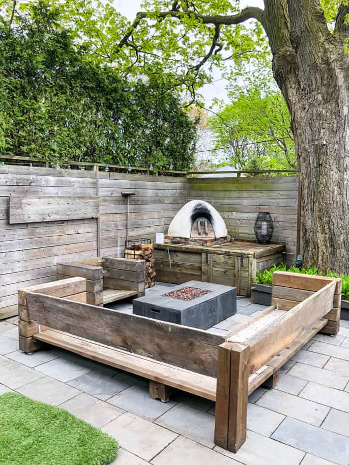 DIY outdoor wooden Restoration Hardware sectional sofa without cushions in front of unlit outdoor fire pit and pizza oven. Wooden fence and large tree in background. 