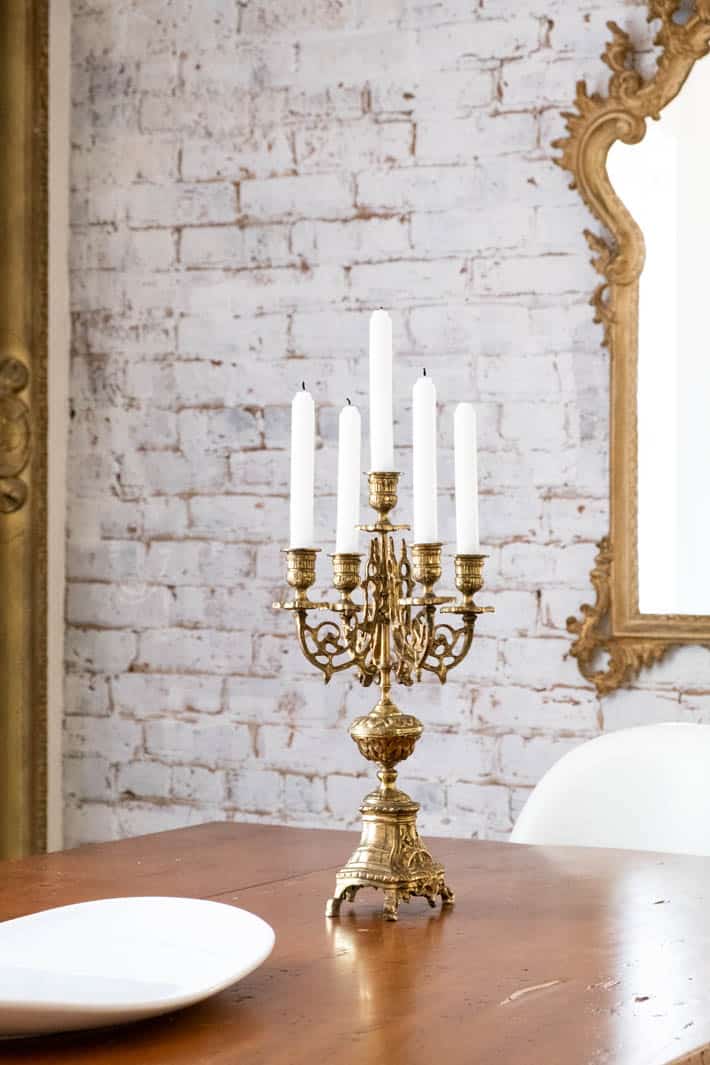 Antique brass candelabra with unlit candles on wooden farmhouse table in front of white painted brick wall in Karen Bertelsen's historic dining room. 