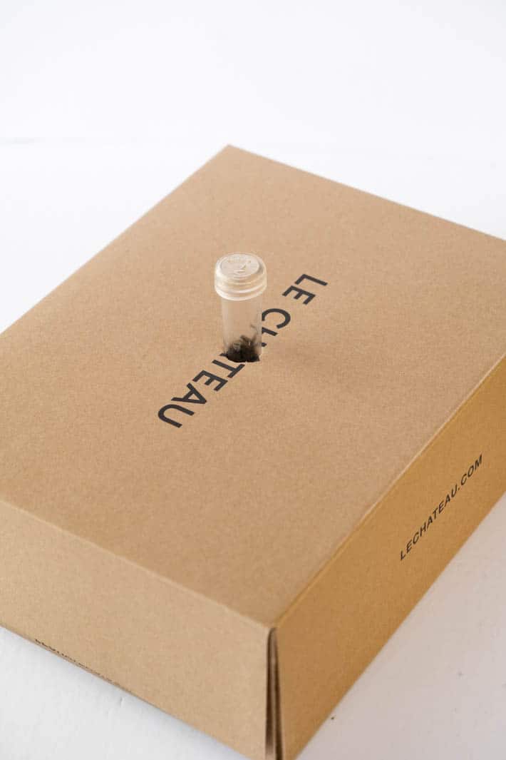 Brown Le Chateau box with translucent floral vial inserted into hole in center of box on white background. 