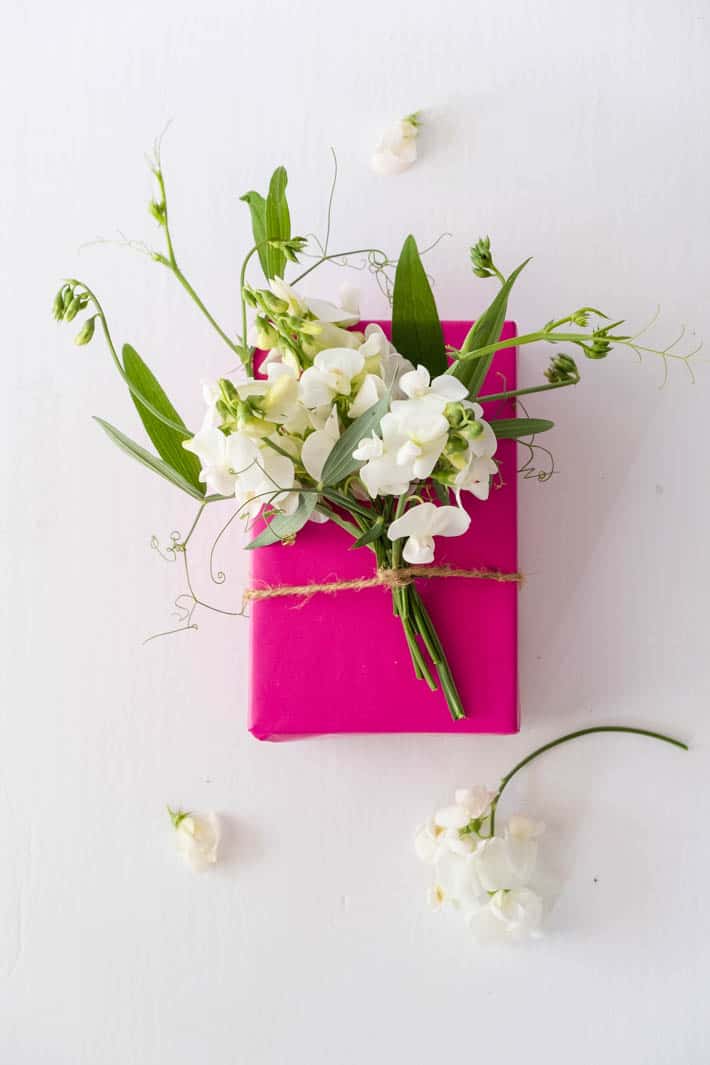 Overhead shot of gift wrapped with fuchsia paper topped with flower bow containing Perennial Sweet Peas, Sweet Pea tendrils and greenery tied on with twine on white background. Loose sweet pea blooms on table in foreground.