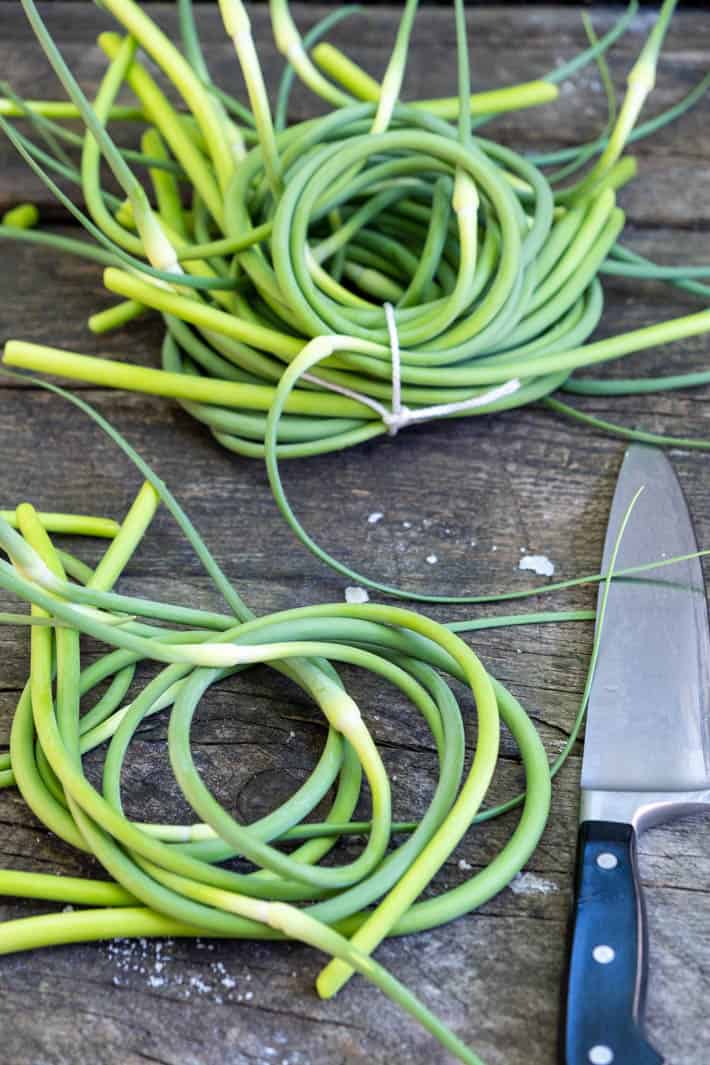 Coils of garlic scapes laid on a rough wood table, tied up with string. A black handled knife set off to the side.