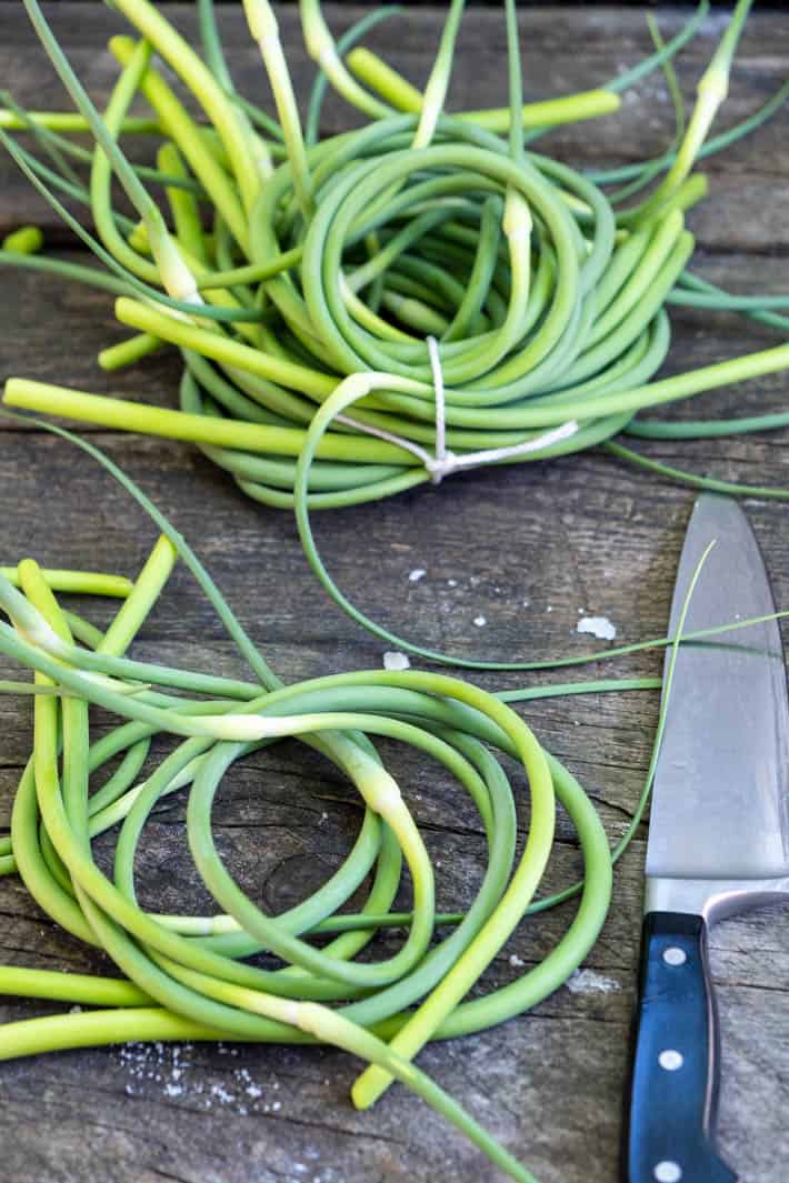 Garlic Scape pesto - the pesto that will ruin you for all other pestos. Coils of garlic scapes laid on a rough wood table, tied up with string. A black handled knife set off to the side.