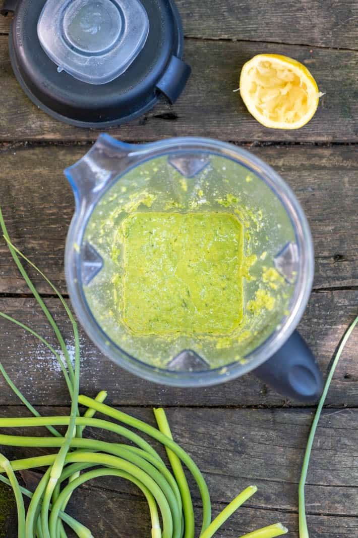 Bright green garlic scape pesto in blender. on a rough wood table with a squeezed lemon half off to the side and garlic scapes coil on the other side.