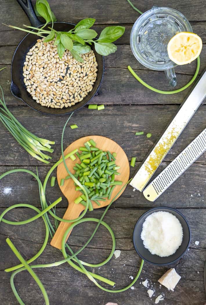 Ingredients for garlic scape pesto laid out on rough wood table. Pine nuts in cast iron pan, lemon juicer and lemon, rasp with lemon zest, grated parmesan cheese in small black bowl, chopped scapes on small wood cutting board and full scapes swirling around.