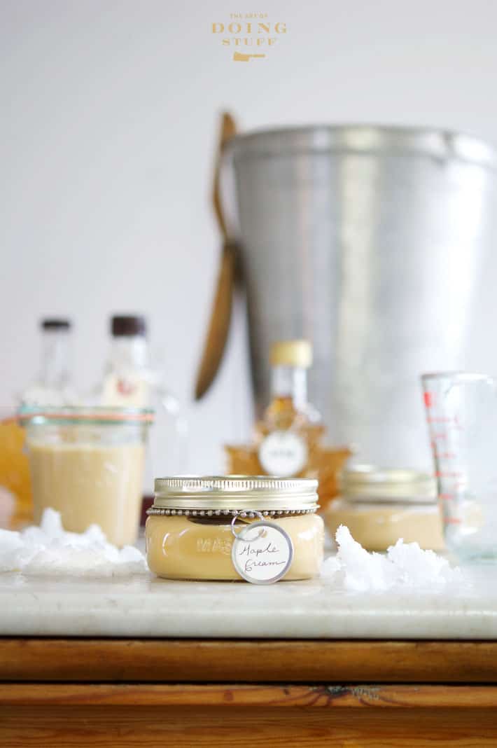 Display of mason jars filled with maple cream and bottles of maple syrup on an antique pine counter with a marble top. Sap bucket with wood spoon in background.