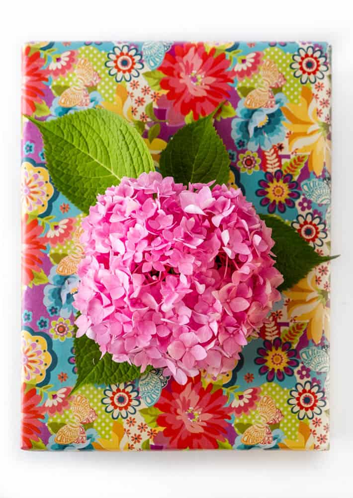 Top-down view of gift wrapped with floral paper topped with pink hydrangea "bow" on white background. 