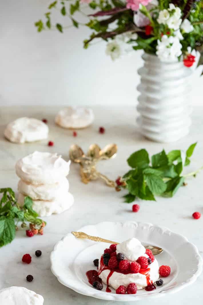 Meringue topped with raspberry compote, fresh berries and whipped cream on white plate with silver spoon. Vase of flowers in background. 