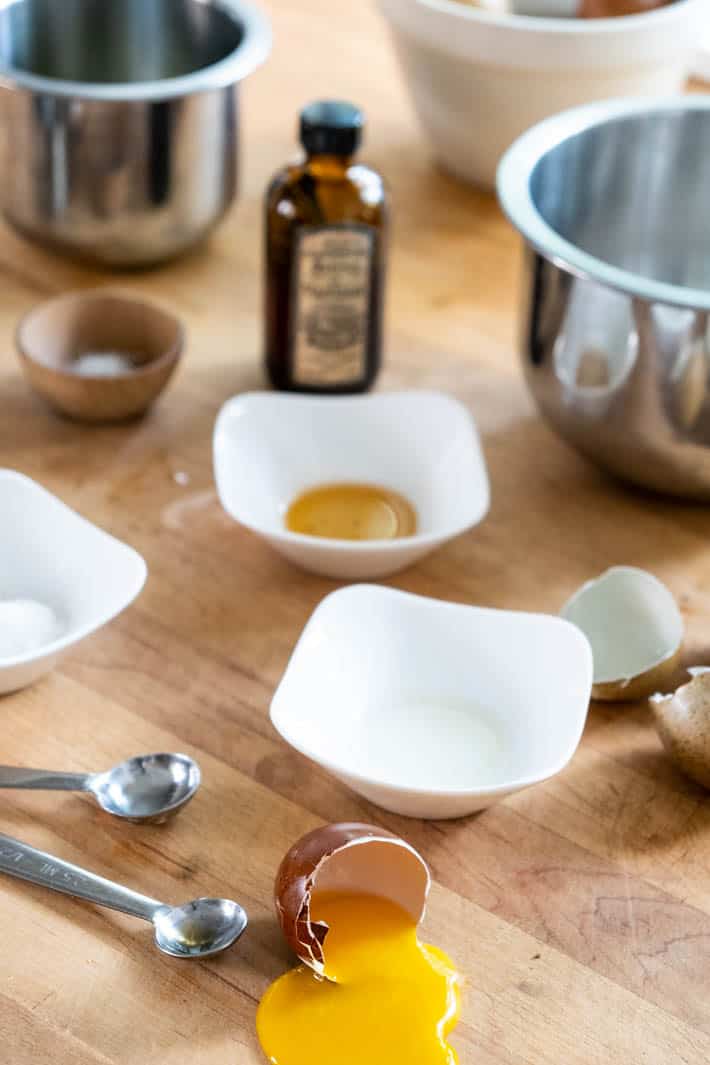 Cracked eggshells, silver measuring spoons, bottle of vanilla extract and white and stainless-steel bowls on wooden countertop. 