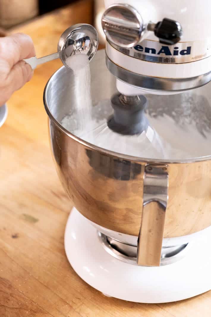 Hand adding a tablespoon of sugar to mixing bowl containing beaten egg whites. 