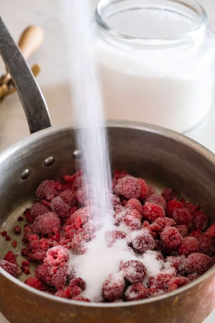 Pouring sugar into pot containing frozen raspberries. 