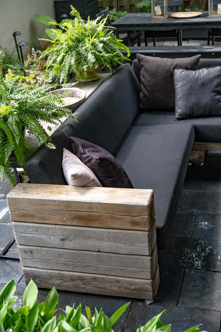 DIY outdoor wooden Restoration Hardware sectional sofa with cushions in place beside potted ferns on table behind sofa.