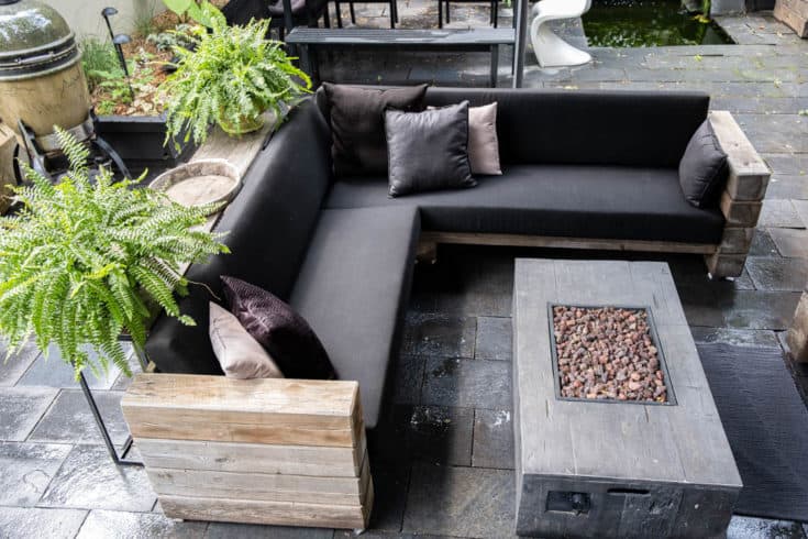 Overhead view of DIY outdoor wooden Restoration Hardware sectional sofa with cushions in place beside unlit outdoor fire pit. Potted ferns sitting on table behind sofa. 