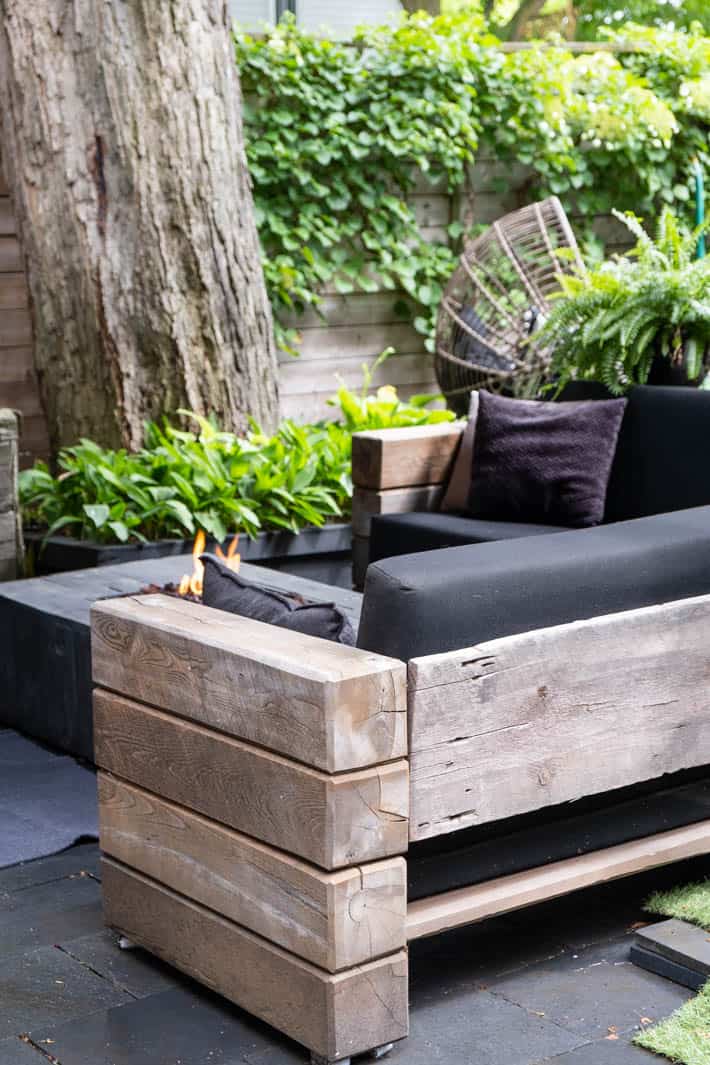 DIY outdoor wooden Restoration Hardware sectional sofa with cushions in front of lit backyard fire pit with large tree and wooden fence covered in greenery in background. 