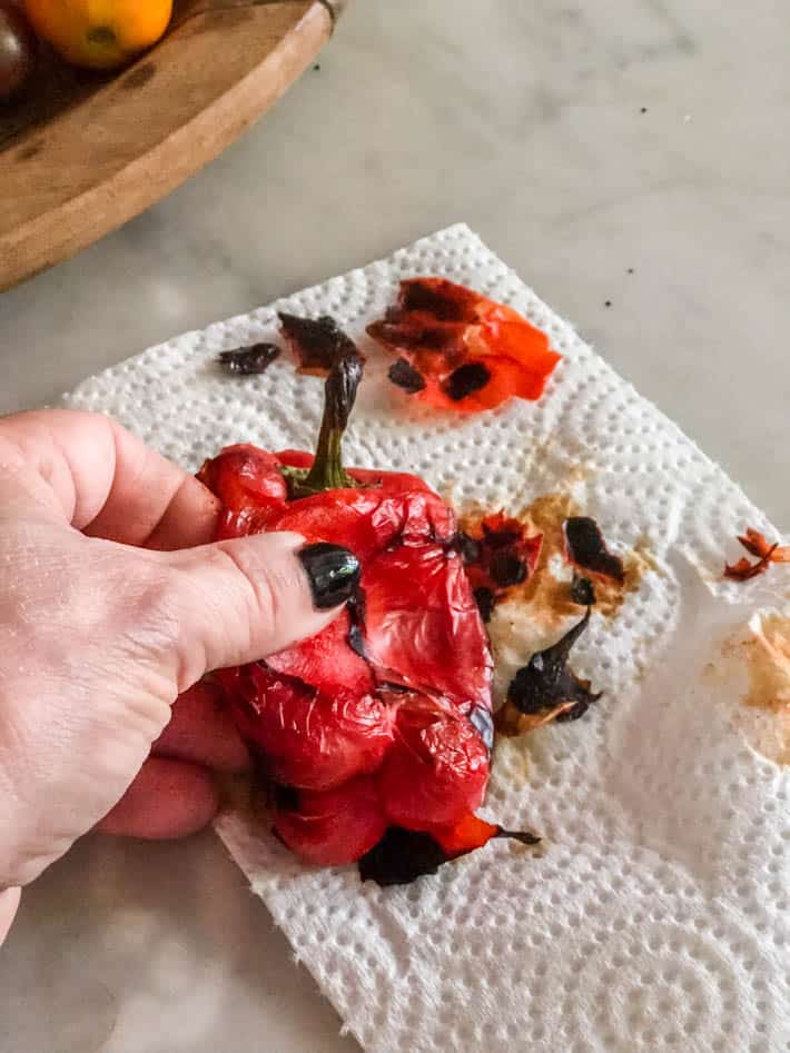 A woman's hand holds a charred roasted red pepper on a white paper towel sitting on a marble kitchen counter. Part of a wooden bowl containing tomatoes can be seen in the background. 