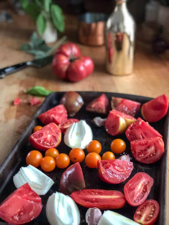 Chopped tomatoes and onion on a dark roasting pan sitting on a butcher block countertop. Tomato, knife, herbs and silver bottle in background. 