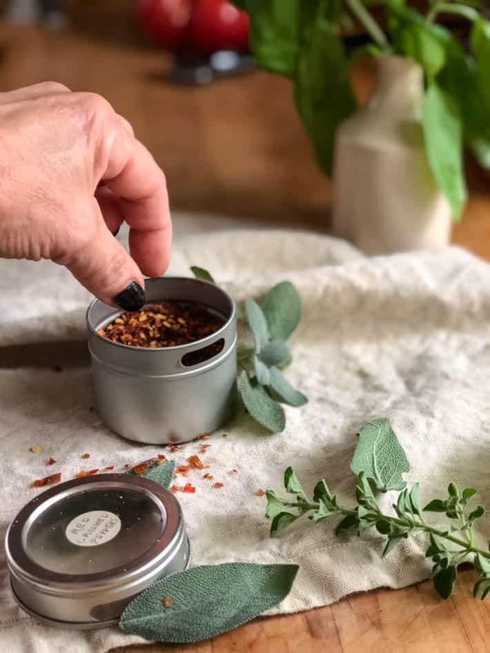 A woman's hand reaches for a tin container of dried red chili flakes sitting on a beige linen tea towel. Fresh sage and oregano seen in foreground. Background shows an ironstone jar containing fresh basil on a butcher block countertop. 