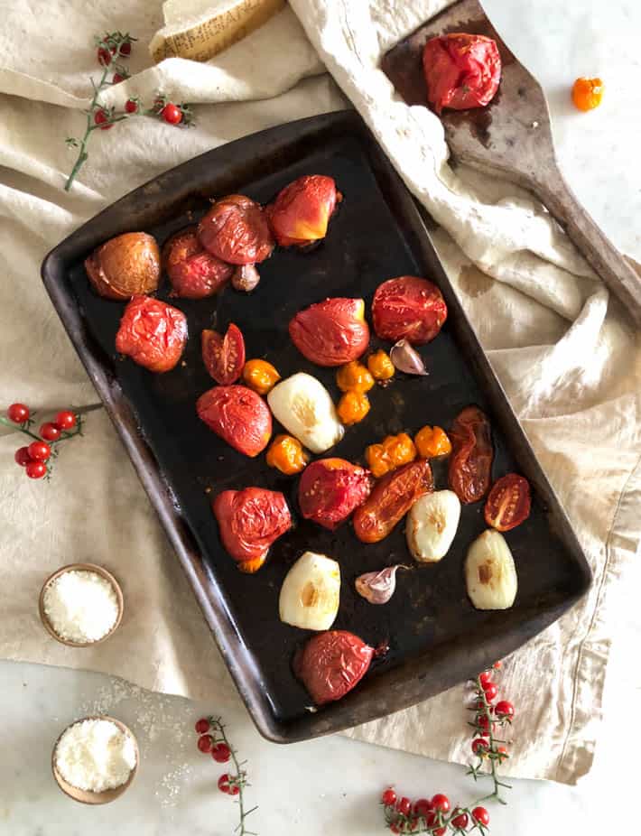 Roasted heirloom tomatoes and onions on a dark baking tray sitting on a beige linen tea towel. Also on towel are tomato vines, a wooden spoon with a roasted tomato and two small wooden bowls containing Parmesan cheese. 