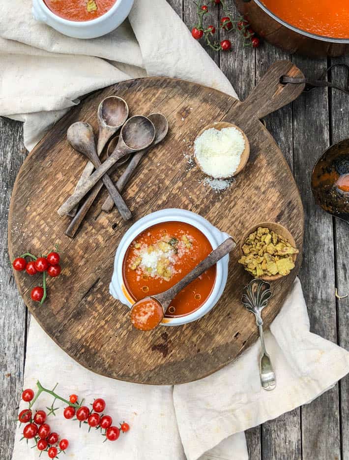 A white soup bowl containing heirloom tomato soup sits on a rustic wooden cutting board on a beige linen tea towel. Also on the cutting board are wooden spoons, two small wooden bowls containing Parmesan cheese and croutons, a tomato vine and a silver spoon. Part of a bowl and a pot of tomato soup can be seen in the background. 