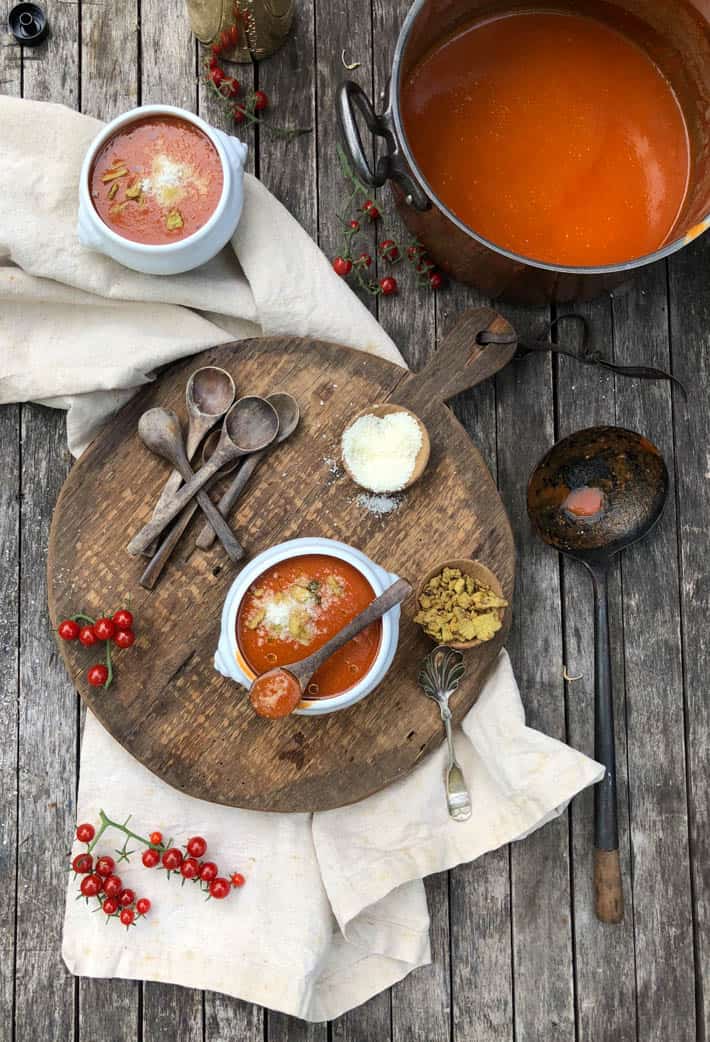 A white bowl containing heirloom tomato soup sits on a wooden cutting board on a beige linen tea towel. In the background are another white bowl and a copper pot both containing heirloom tomato soup.