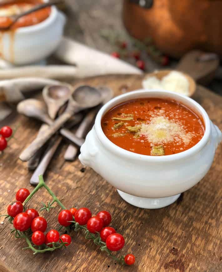 A white soup bowl containing heirloom tomato soup sprinkled with Parmesan cheese sits on a rustic wooden cutting board. Also on board are wooden spoons and tomato vines. A copper pot and another bowl of heirloom tomato soup are seen in background. 