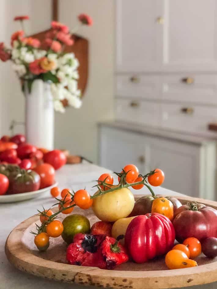 A shallow wooden bowl containing various heirloom tomatoes and a roasted red pepper can be seen in the foreground. The background shows tomatoes on a white plate, a white vase with flowers and white kitchen cupboards. 