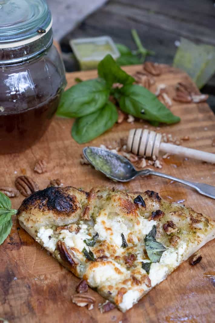 A slice of pesto pizza on wooden cutting board in foreground. Spoon covered with pesto, wooden honey dipper, fresh basil, pecans, containers of pesto and honey pot can be seen in background. 