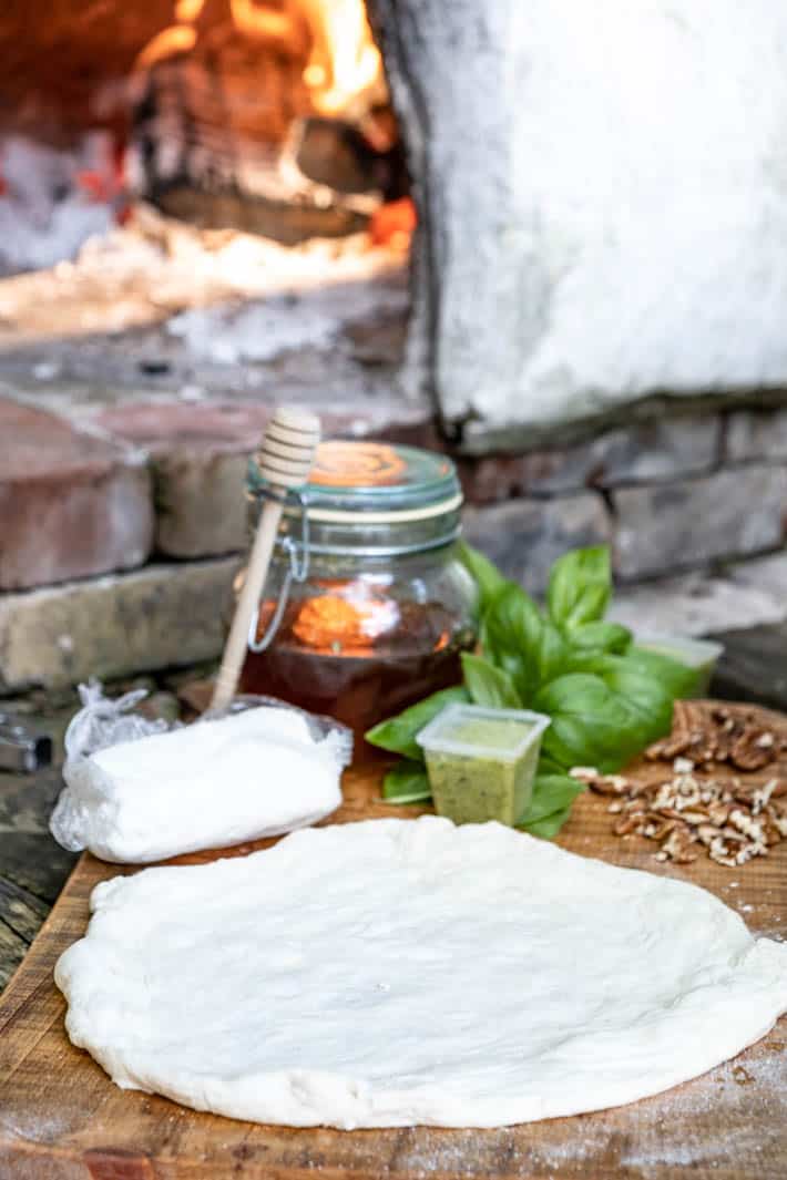 Disc of uncooked pizza dough, goat cheese, garlic scape pesto, pecans, fresh basil and a honey pot with honey dipper on wooden cutting board. Pizza oven with burning logs in it in background. 