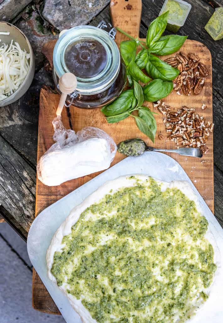 Overhead view of rolled out dough slathered with pesto on wooden cutting board. Also on board are goat cheese, chopped and whole pecans, bunches of fresh basil, and honey pot with honey dipper. Shredded mozzarella and containers of garlic scape pesto in background. 