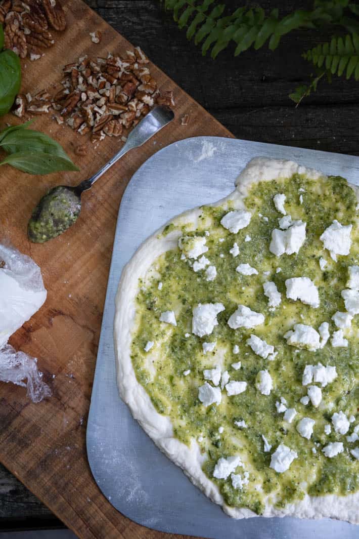 Rolled out pizza dough covered with garlic scape pesto and chunks of goat cheese on a wooden cutting board. Also on cutting board are a spoon covered with pesto, pecans, fresh basil and log of goat cheese. 