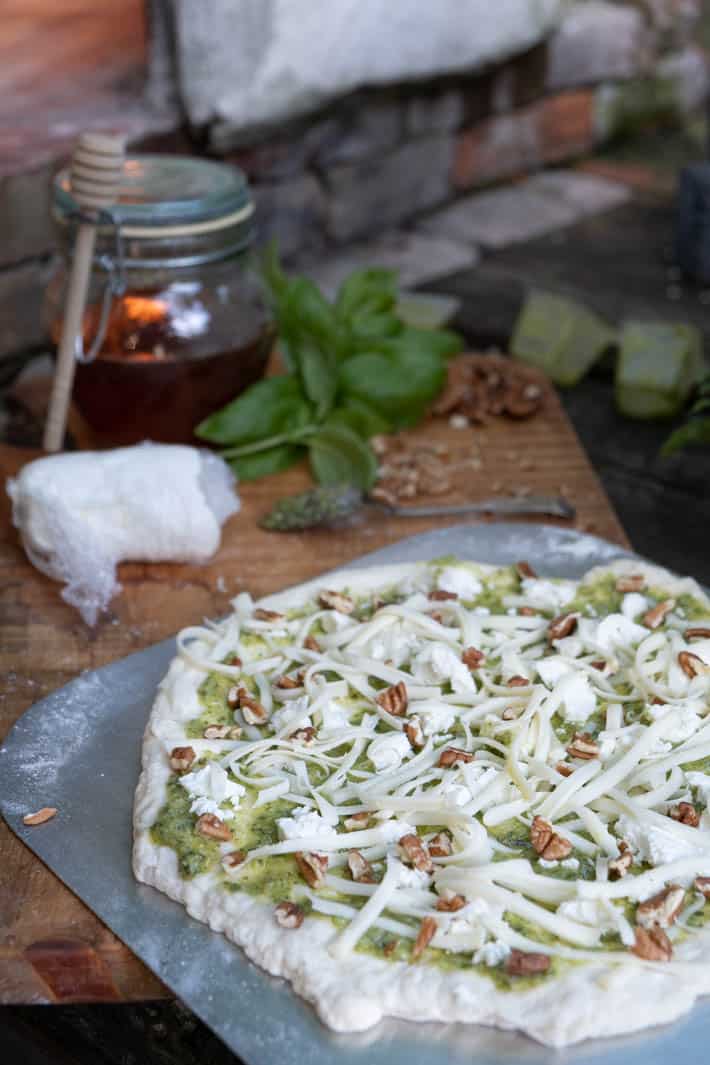 Uncooked pizza dough covered with pesto, chunks of goat cheese, mozzarella and chopped pecans on wooden cutting board in front of pizza oven. 