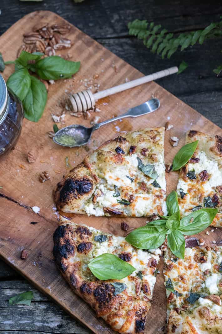 Freshly baked and cut pesto pizza on a wooden cutting board with a silver spoon, honey dipper and pot of honey in background. 