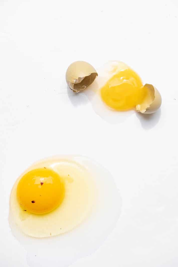 Two cracked eggs (one with the broken shell beside it) on a white background. The yolk of the egg in the foreground has two blood spots.