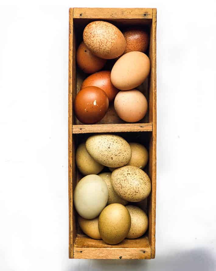 Eggs in various shades of brown and pale-olive in a wooden crate on a white background. 