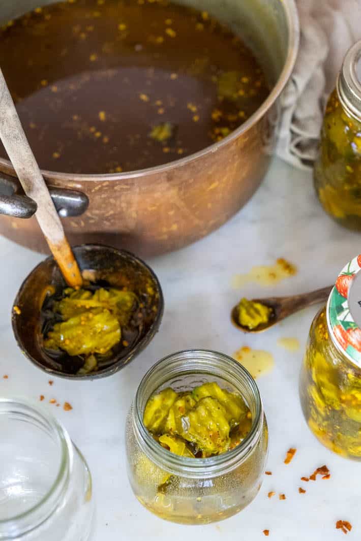 A copper pot containing pickling liquid sits on a marble counter top with a copper ladle and clear glass jars filled with pickles around it. 