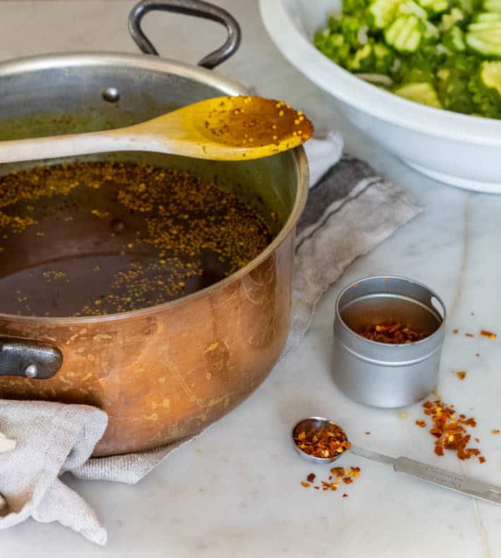 A copper pot filled with canning liquid sits on a marble counter top. Beside the pot is a small spice jar containing red chili flakes. Sliced cucumbers in a white bowl can be seen in the background. 