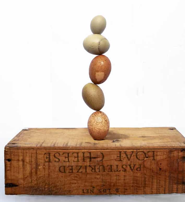Eggs stacked on top of each other on a rustic wooden crate against a white background. At the top of the stack is a fart (or fairy) egg. 
