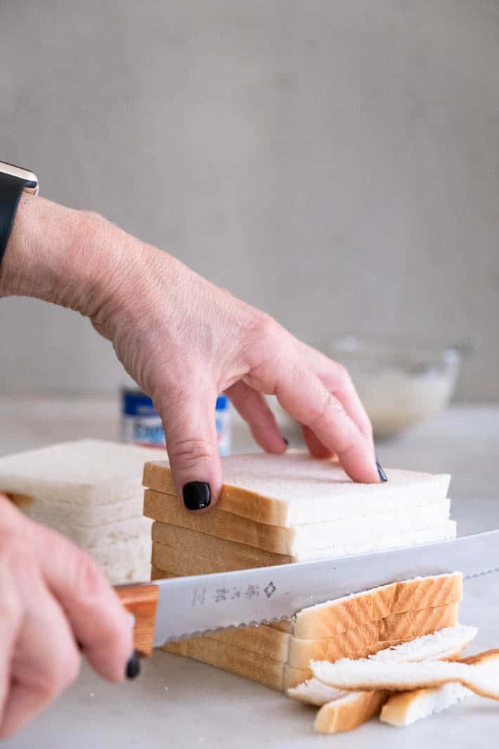 A stack of soft white bread with the crusts being cut off with a wood handled serrated knife.