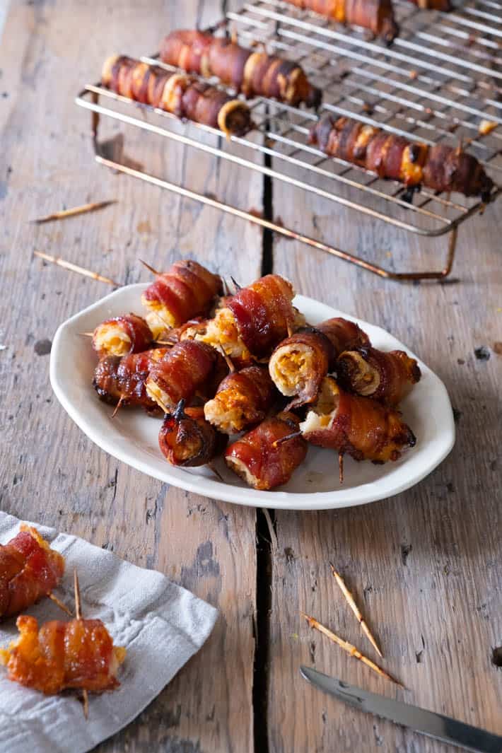 Cooked bacon wraps with toothpicks on an ironstone plate with a cookie cooling rack in the background.
