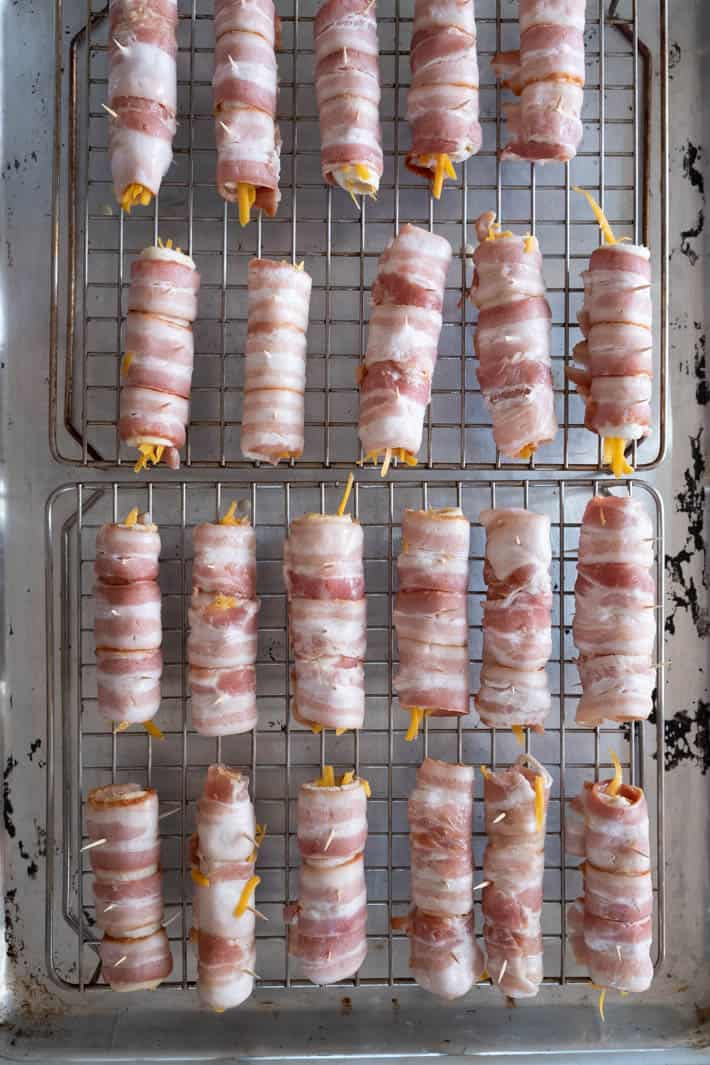 Crispy, gooey bacon wraps with a perfect combination of salt and a tiny touch of sweet. #bacon #appetizers Rows of uncooked bacon wraps on cookie racks set on well used baking sheet.