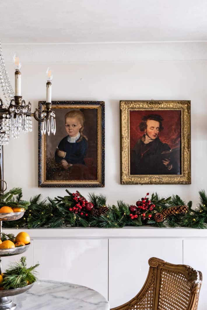 Two vintage oil portraits in gold frames, side by side over modern white lacquer buffet with lush Christmas garland with red berries.