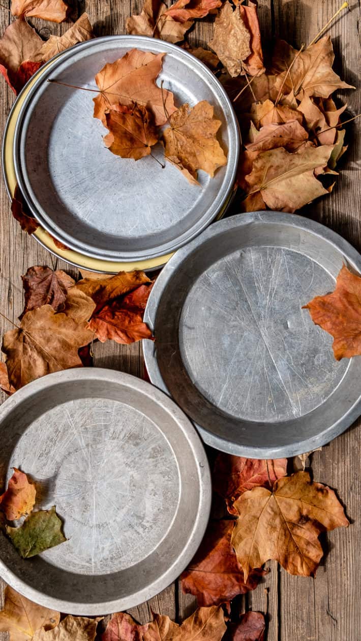 3 old aluminum pie plates on a wood table with scattered maple leaves around.