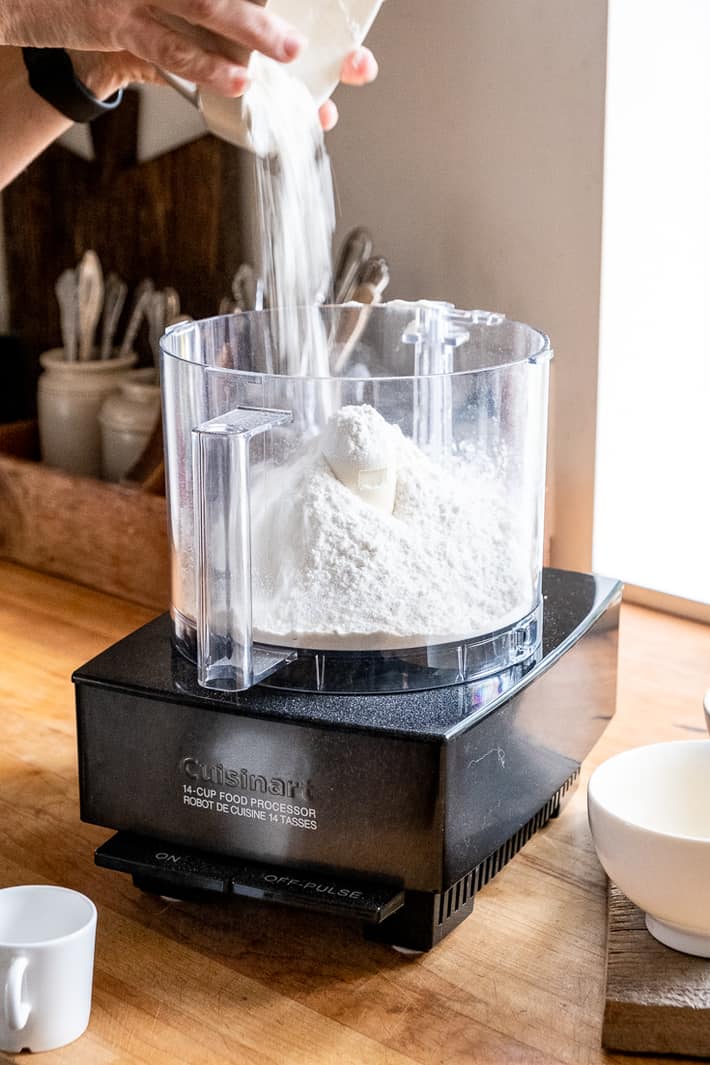 Flour being poured out of an ironstone bowl into a food processor on a butcher block countertop.