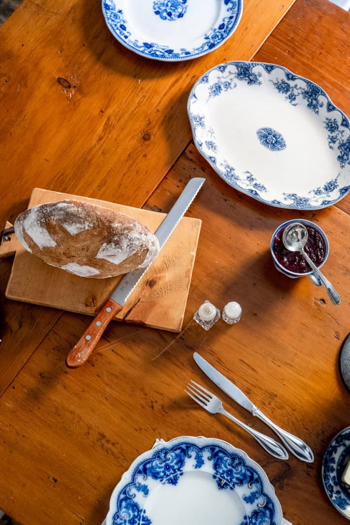 Freshly baked bread sits on a cutting board with a serrated knife on a harvest table with flow blue dishes around.
