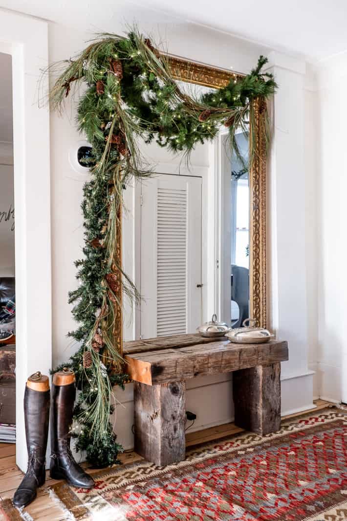 Mixed artificial garland swags over a huge ornate gold mirror on the wall, with riding boots and a bench beneath.