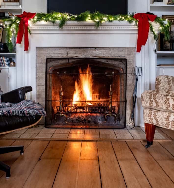 A white mantled fireplace, with a fire roaring in the hearth and evergreen branches with red bows on the mantle.