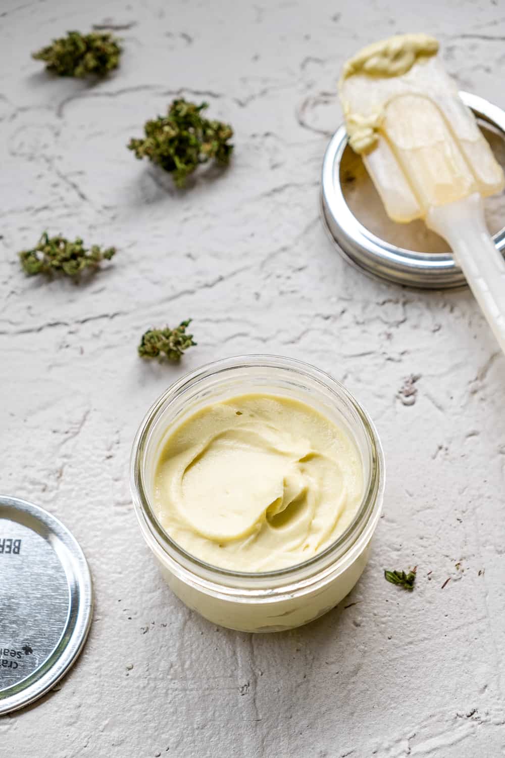 Whipped cannabutter in a small, low, jam sized mason jar with cannabis scattered around and spatula off to the side.